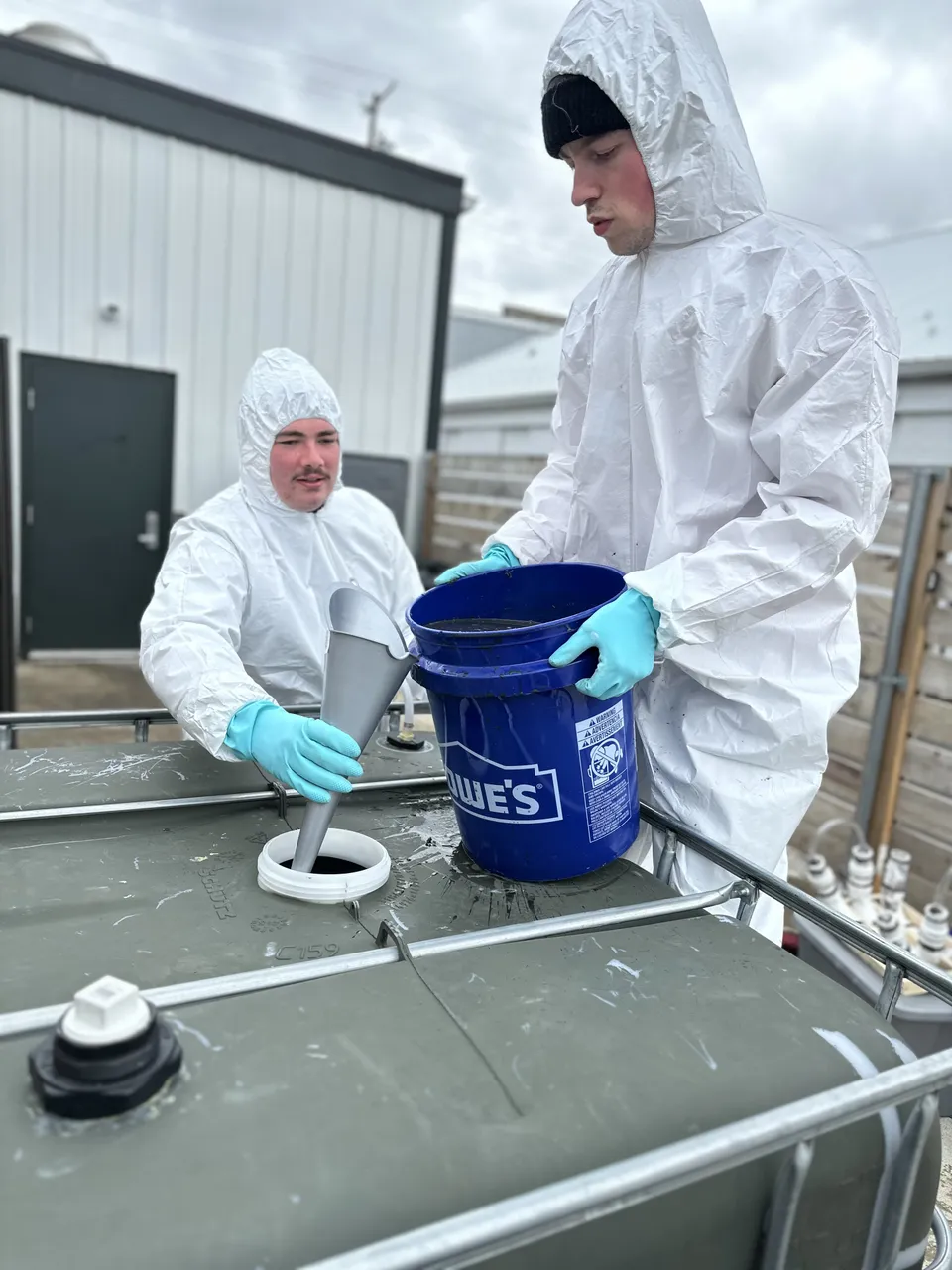 Two EvE Waste team members in white protective suits loading feedstock from a bucket through a funnel into the AD system prototype.